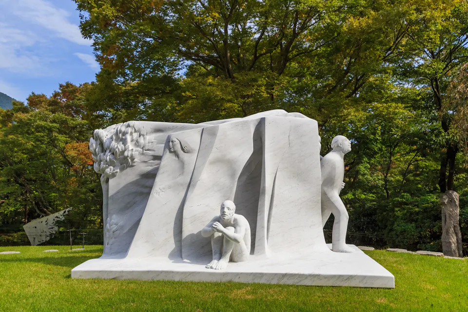 白い大理石の彫刻が緑の草の上に立ち、木々の背景に対峙しています。このアートワークは、三人の人間の姿を描いています：一人は考え込んで地面に座り、もう一人は壁から部分的に現れ、三人目は構造物から離れて歩いています。