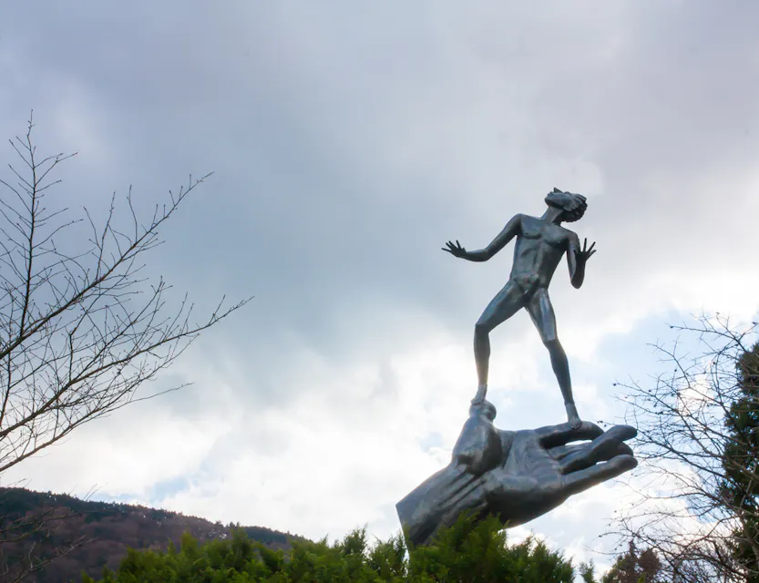 空に向かって手を伸ばす金属製の手の上に人間の姿をした像が立っています。背景には、一部雲がかかった空と、前景には丘と裸の木の枝があります。このシーンは、向上心と希望の感覚を伝えています。