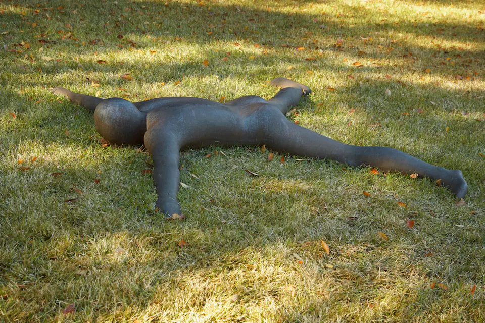 草地の上に手足を広げてうつ伏せに寝る人間の姿の青銅の彫刻です。この像は実物大に見え、リラックスした大の字の姿勢で、散らばった葉に囲まれています。