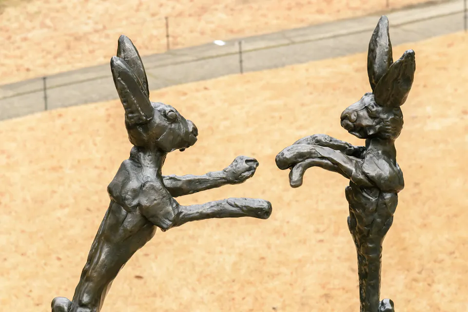 2匹の人間のようなウサギの像が後ろ足で立って遊び心満載のボクシングのポーズをとっている。背景には砂色の表面があり、遠くにぼやけた小道が見える。