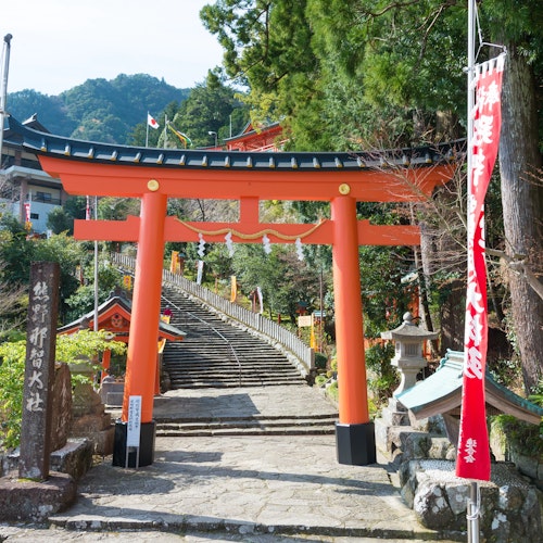 Pilgrimage Routes and Spiritual Treks in Japan A traditional Japanese torii gate stands at the entrance of a shrine, with steps leading up behind it. Red banners with white text flank the gate, and lush greenery surrounds the area. A red shrine building is visible in the background.