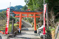 A traditional Japanese torii gate stands at the entrance of a shrine, with steps leading up behind it. Red banners with white text flank the gate, and lush greenery surrounds the area. A red shrine building is visible in the background.
