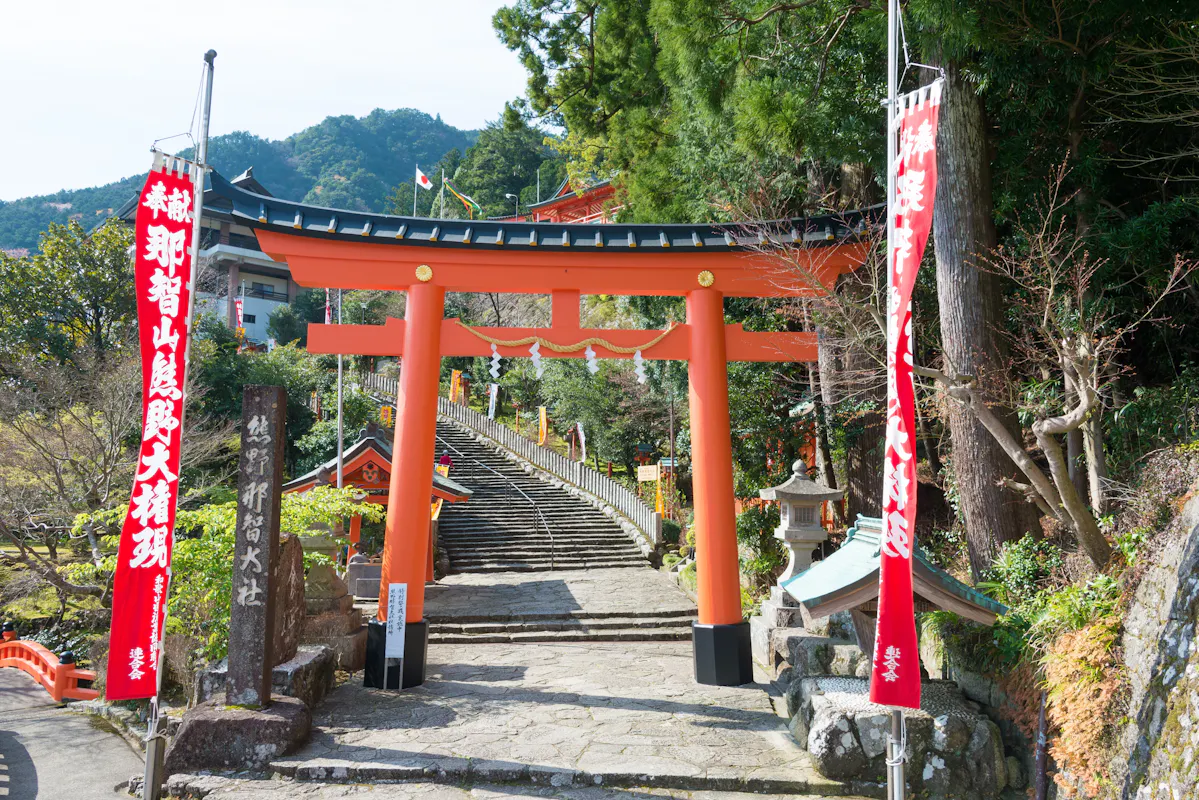 A traditional Japanese torii gate stands at the entrance of a shrine, with steps leading up behind it. Red banners with white text flank the gate, and lush greenery surrounds the area. A red shrine building is visible in the background.