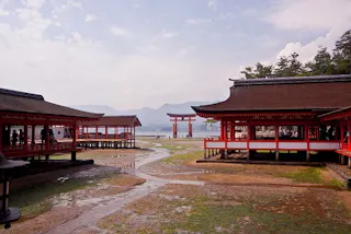 Traditional Japanese Shinto shrine buildings with red columns and thatched roofs stand near the shoreline, with the iconic floating torii gate visible in the distance, surrounded by mountains and a cloudy sky.