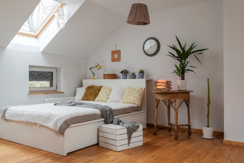 A cozy bedroom with a slanted ceiling and a skylight. The room has a white bed with patterned yellow pillows and a gray throw. A wicker table with candles and plants is to the right, while a crate and a gray blanket lie at the foot of the bed. A wall clock and a pot plant are in the background.