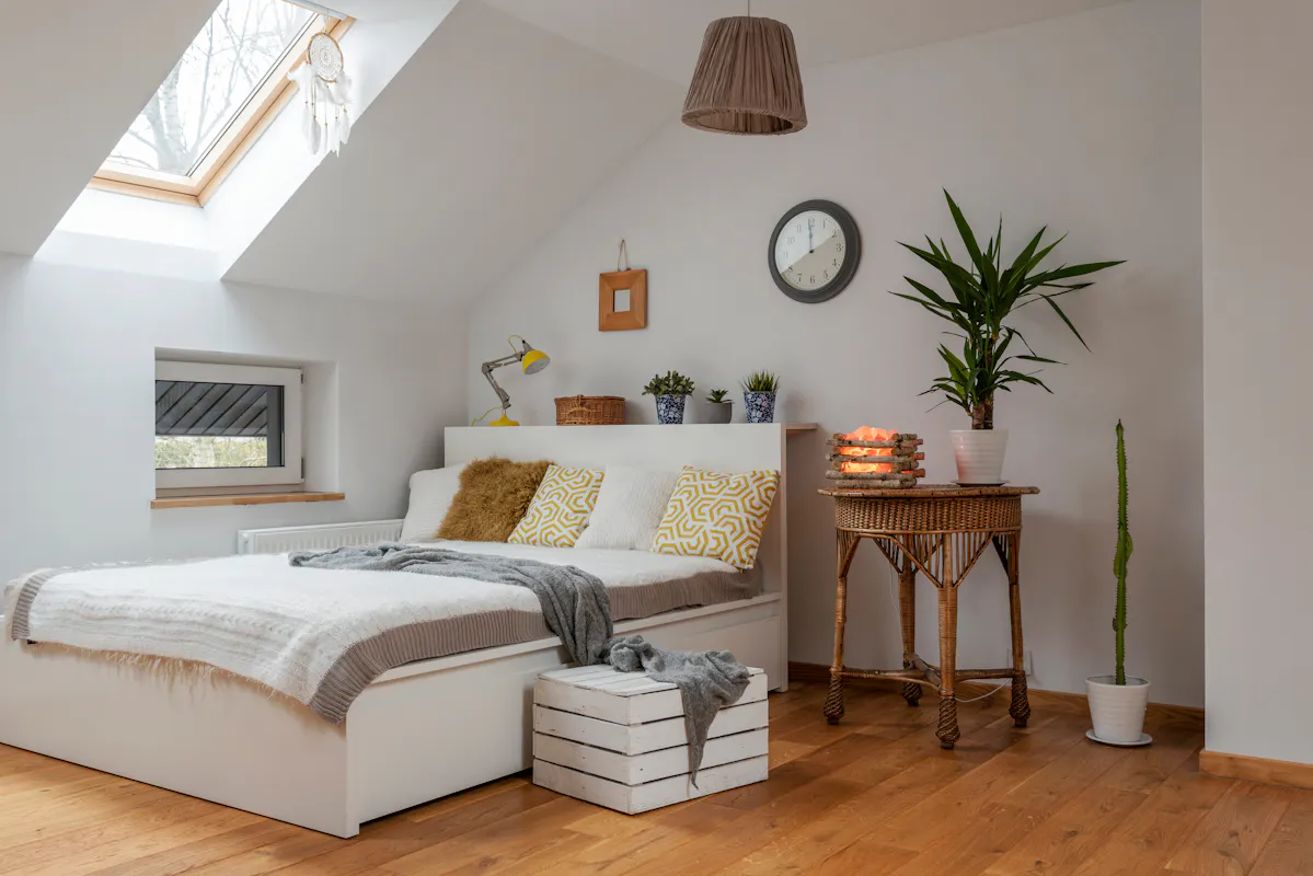 A cozy bedroom with a slanted ceiling and a skylight. The room has a white bed with patterned yellow pillows and a gray throw. A wicker table with candles and plants is to the right, while a crate and a gray blanket lie at the foot of the bed. A wall clock and a pot plant are in the background.