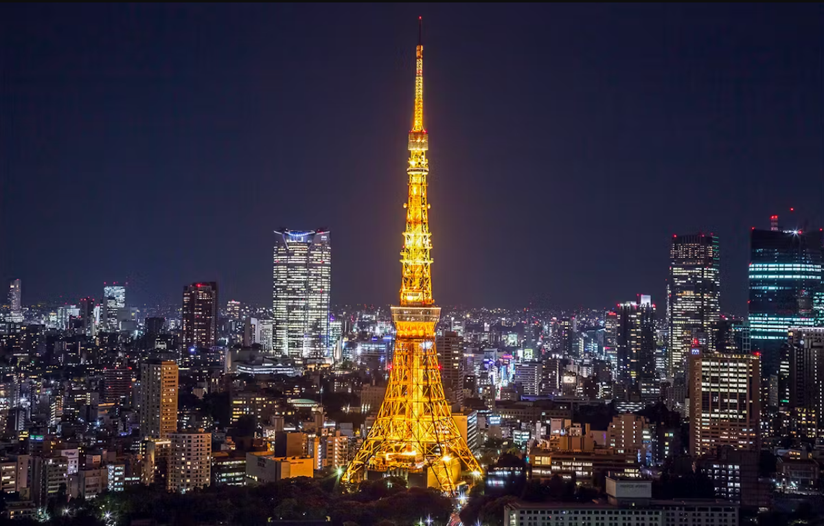 A night view of a city with a brightly illuminated tower in the center, resembling Tokyo Tower. Surrounding the tower are numerous buildings with scattered lights, creating a vibrant cityscape against a dark sky.
