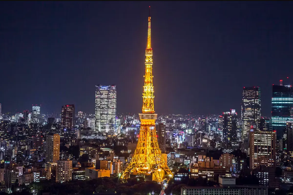 Tokyo Tower A night view of a city with a brightly illuminated tower in the center, resembling Tokyo Tower. Surrounding the tower are numerous buildings with scattered lights, creating a vibrant cityscape against a dark sky.
