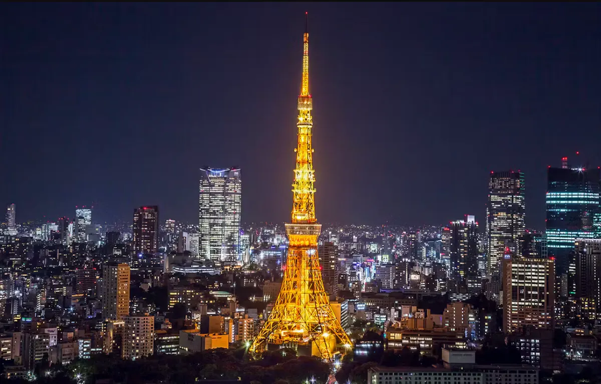A night view of a city with a brightly illuminated tower in the center, resembling Tokyo Tower. Surrounding the tower are numerous buildings with scattered lights, creating a vibrant cityscape against a dark sky.