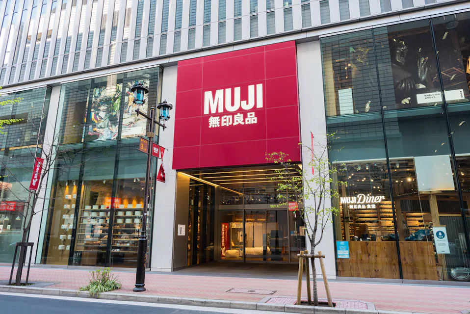 Muji Hotel Ginza Street view of the front entrance of a MUJI store with large red signage displaying the MUJI logo and Japanese characters. The store has glass windows, showcasing well-lit interiors with various products and a MUJI Diner on the right side. Small trees and street lamps are along the sidewalk.