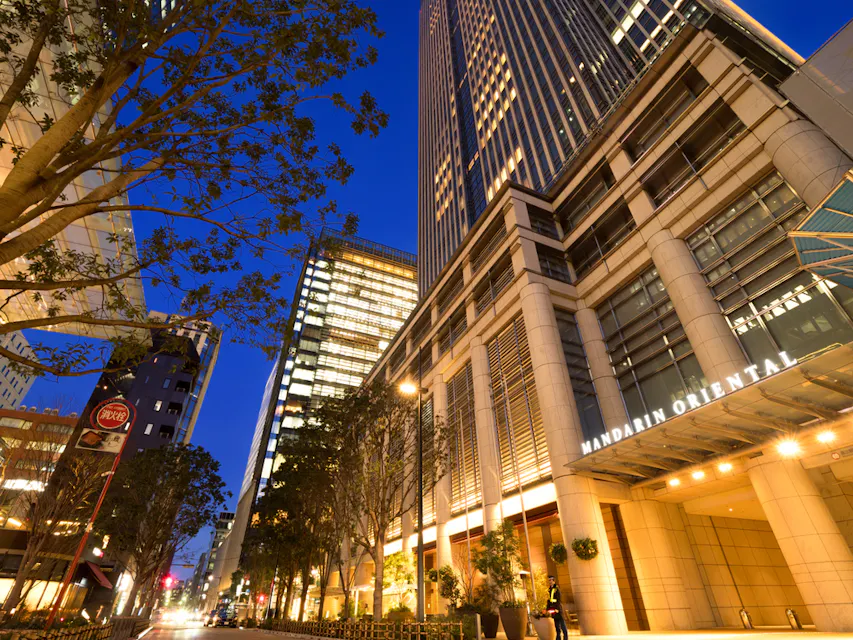 Mandarin Oriental Tokyo A vibrant city street at dusk features tall buildings, with one prominently marked "Mandarin Oriental" on its façade. Trees line the sidewalks, and the building windows are brightly lit, contrasting with the deep blue sky.