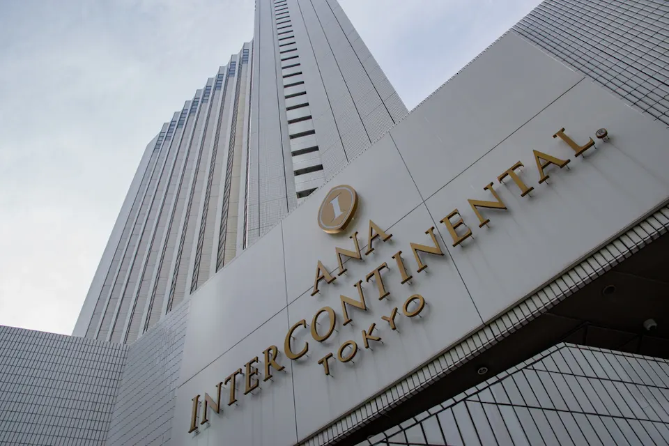ANA InterContinental Tokyo A low-angle shot of the ANA InterContinental Tokyo, featuring a tall, modern skyscraper. The building's facade has large gold letters spelling "ANA InterContinental Tokyo" with a round logo above it. The sky appears cloudy in the background.