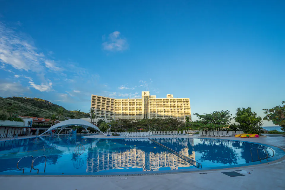 Renaissance Okinawa Resort A large hotel with a prominent "R" sign on the roof stands behind a spacious swimming pool under a clear blue sky. The pool has a decorative arch, lounge chairs arranged in rows, and a backdrop of green hills and trees.