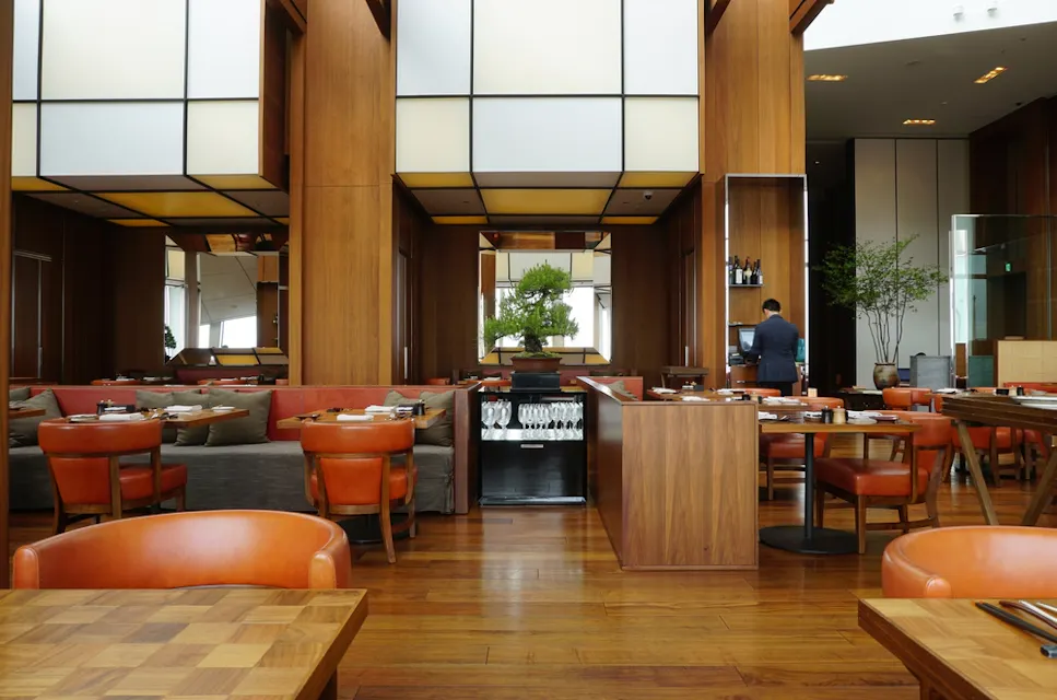 Andaz Tokyo – A Concept by Hyatt A spacious, modern restaurant with wooden floors and decor. It features orange leather chairs and wooden tables neatly arranged. A person stands in the background near the side, and potted trees add greenery. The ambiance is bright and inviting.