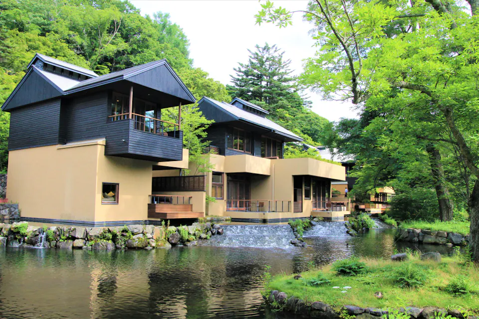 Hoshinoya Karuizawa A serene scene featuring modern houses with black and beige exteriors situated beside a tranquil pond and a small cascading waterfall. Lush green trees surround the buildings, reflecting a peaceful, natural setting in the water below.