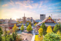A panoramic view of Senso-ji Temple in Tokyo, Japan, with its iconic pagoda and ornate main hall surrounded by trees. People stroll through the temple grounds on a sunny day, with modern city buildings visible in the background.