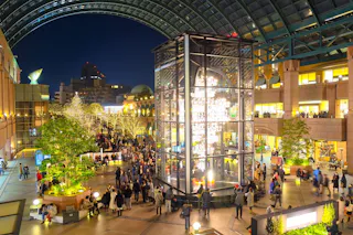A bustling indoor plaza at night, decorated with festive lights and a large illuminated glass tower centerpiece. Crowds of people are walking and gathering in the open space, surrounded by shops, greenery, and trees adorned with string lights. A glass roof arches overhead.