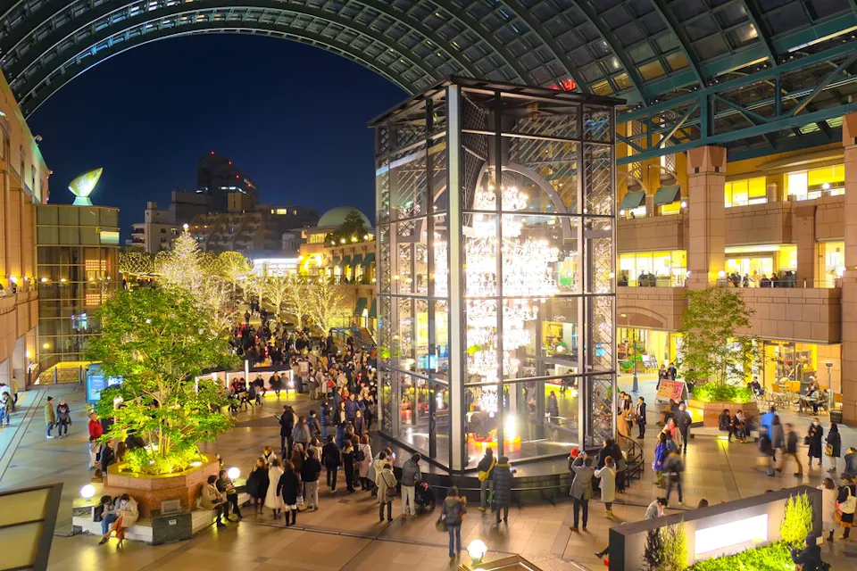 一个热闹的室内广场，夜晚被节日灯光装饰，中央有一个大型的发光玻璃塔。人们在开放空间里走动和聚集，周围是商店、绿树和挂着串灯的树木。弧形的玻璃屋顶高高悬挂在空中。