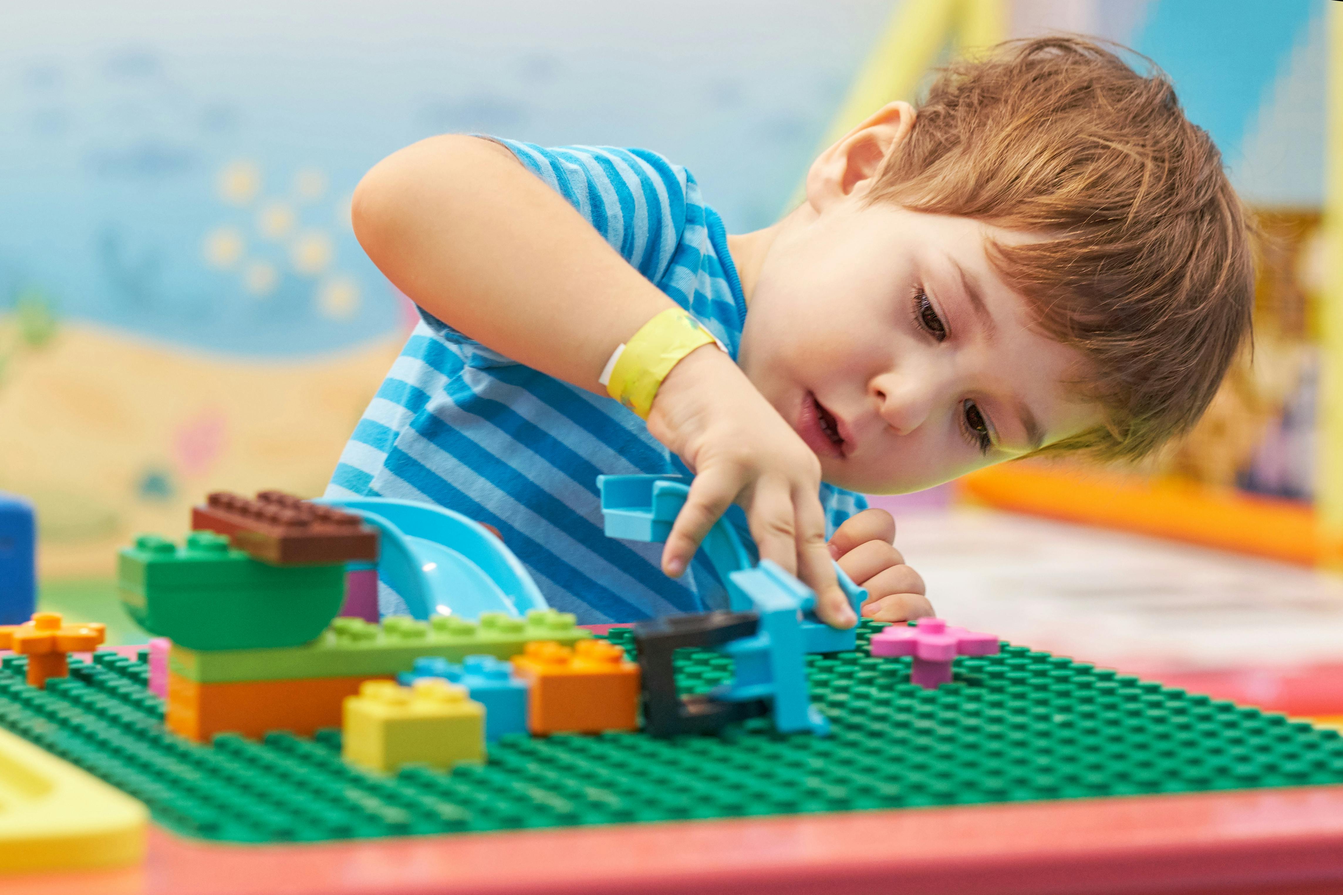 A young child in a blue striped shirt concentrates while building with colorful plastic blocks on a green base plate at a play table.