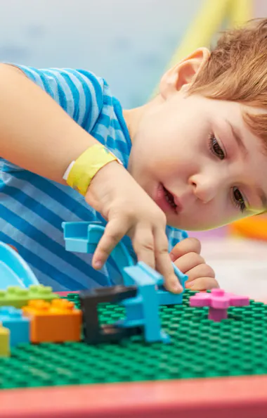 Boy playing with Lego A young child in a blue striped shirt concentrates while building with colorful plastic blocks on a green base plate at a play table.