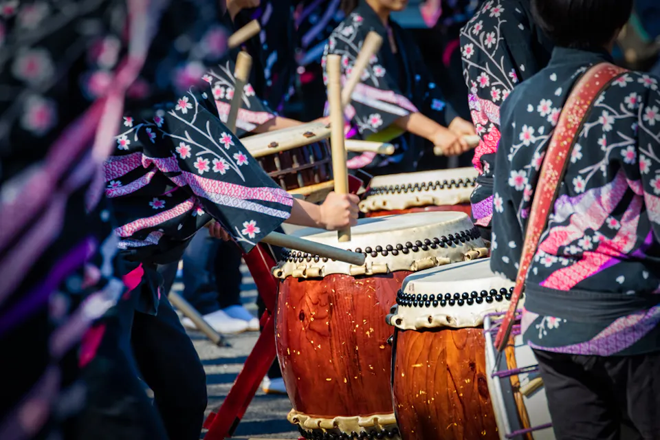 Close-up of people wearing black floral-patterned attire playing traditional Japanese taiko drums. The vibrant image features focused drummers energetically striking the drums with wooden sticks during an outdoor performance.