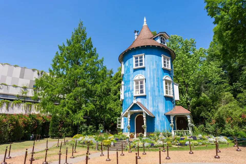 A whimsical, cylindrical blue house with a conical brown roof, surrounded by lush greenery and blooming flowers. The house features small windows and a quaint door. A path with decorative poles leads to the entrance, and the sky is clear and blue.