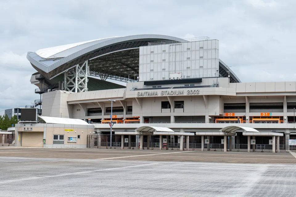 Wide shot of Saitama Stadium 2002, showing its large, modern white structure with a curving roof and multiple entry points. The stadium appears quiet, with no visible crowds, under an overcast sky. The words "SAITAMA STADIUM 2002" are prominently displayed.