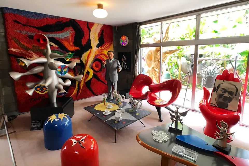 Taro Okamoto Museum of Art A person stands in a creatively decorated room with colorful abstract art. The space features vibrant furniture, unique sculptures, and a large window letting in natural light. The decor includes a red and black tapestry, and a blue sculpture with multiple white arms.