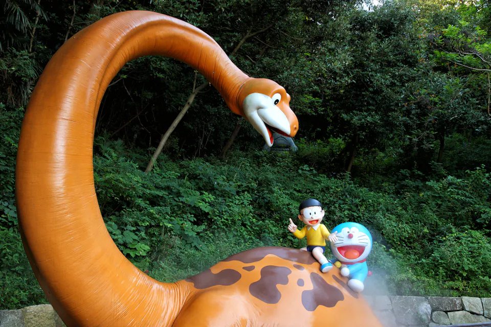 Fujiko F. Fujio Museum A statue featuring a large dinosaur with a long neck looking down at two characters sitting on its back. One character is a boy in a yellow shirt and blue shorts, while the other is a blue robotic cat. They are surrounded by lush greenery.