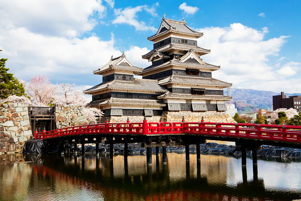 Matsumoto Castle A traditional Japanese castle with layered rooftops stands against a backdrop of mountains and a blue sky. A vibrant red bridge spans a calm moat in the foreground, leading to the castle entrance. Cherry blossoms are visible on the left side.