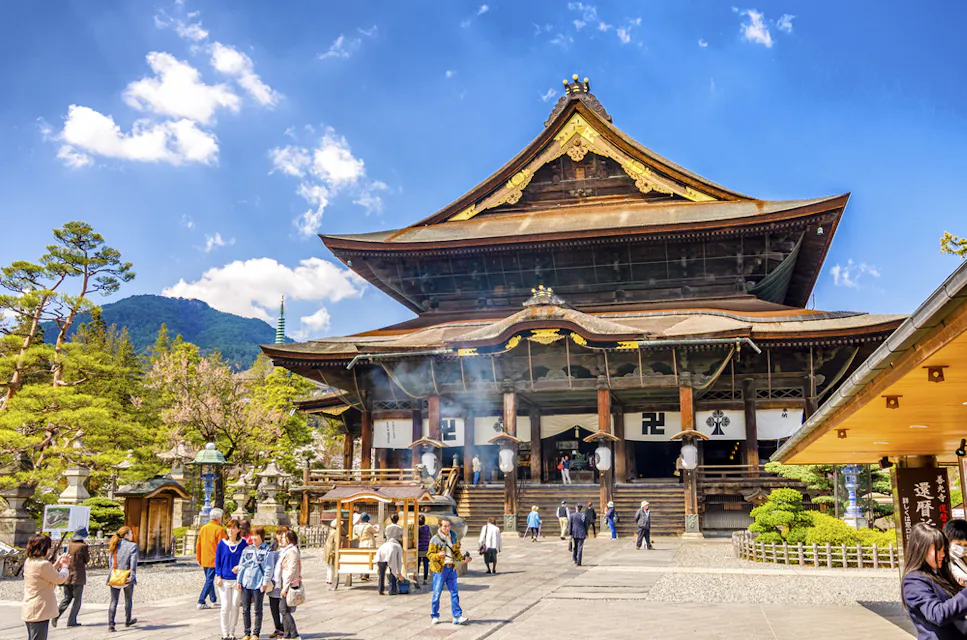 A large traditional Japanese temple with a steep, gabled roof stands under a bright blue sky. Several people are walking around the courtyard in front of the temple. Lush greenery and trees are visible in the background, along with a mountain range.