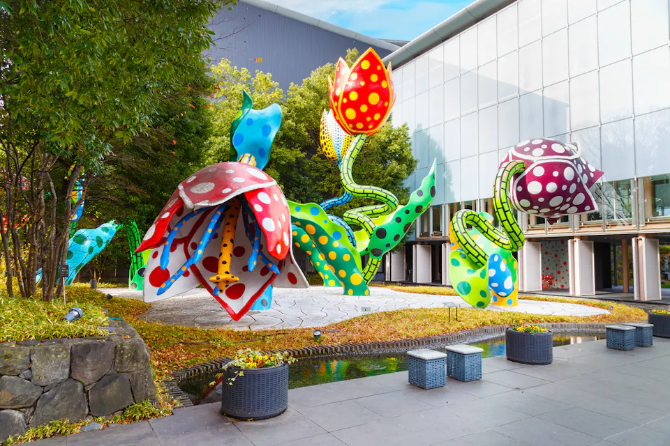 Matsumoto City Museum Colorful, oversized polka-dotted flower sculptures in an outdoor garden setting, surrounded by green foliage and a modern glass building. The sculptures come in various bright colors, like red, blue, yellow, and purple, creating a whimsical and vibrant atmosphere.
