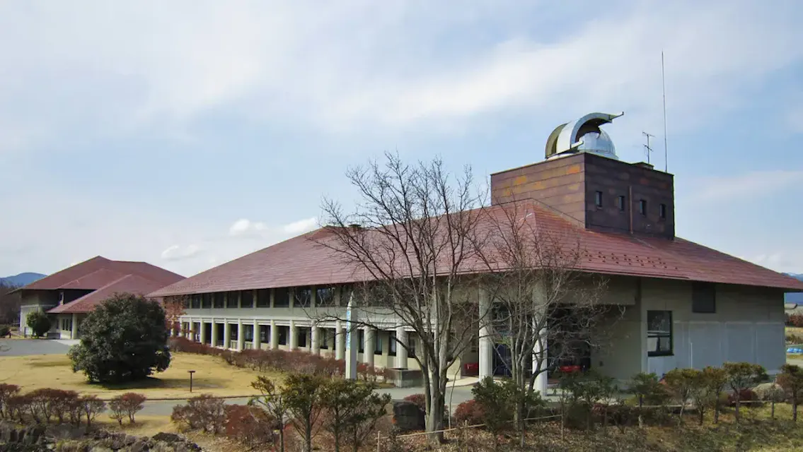 Nagano City Museum A large building with a sloped red roof and a tower featuring a crescent moon sculpture on top. The structure is surrounded by well-maintained bushes and trees, with a clear sky in the background. A smaller building with a similar roof is visible to the left.