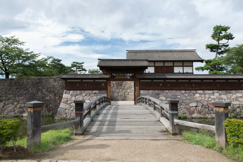 Matsushiro Castle Park A wooden bridge leads to a historic Japanese castle gate, framed by stone walls and surrounded by lush green trees. The sky is partly cloudy, creating a serene atmosphere.