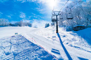 A scenic view of a snow-covered ski slope with a clear blue sky. A ski lift with empty chairs is on the right, casting long shadows on the snow. Trees covered in snow line the edge, and a small mountain hut is visible in the background.