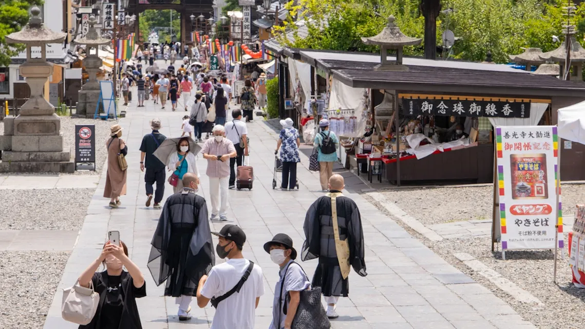 Traveling to Nagano A bustling street scene in Japan, with numerous people walking and engaging with vendors in stalls. Many are wearing masks. Monks in traditional robes are visible. Bright sunlight highlights the green foliage and buildings in the background.