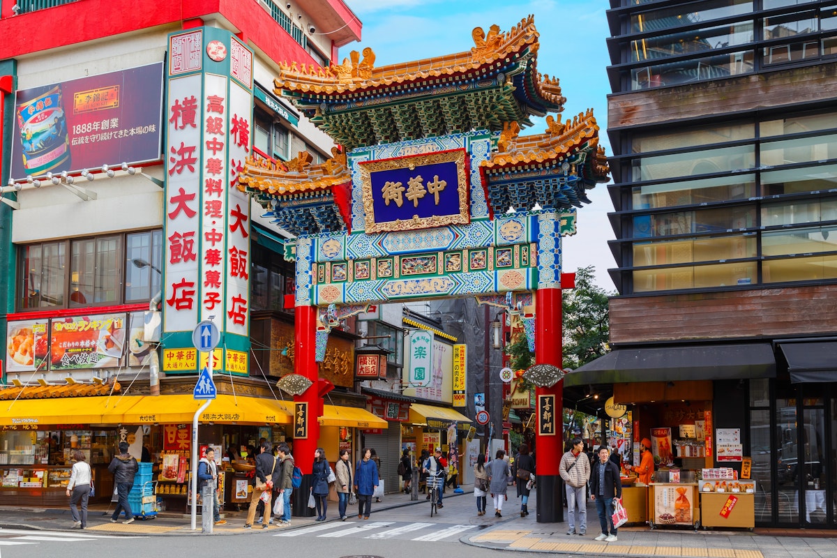 Yokohama's Chinatown Street scene in Yokohama Chinatown, featuring a colorful traditional Chinese gate with intricate designs. People walk and shop at various stores, including restaurants and souvenir shops. Bright signs with Chinese characters are prominently displayed on the buildings.