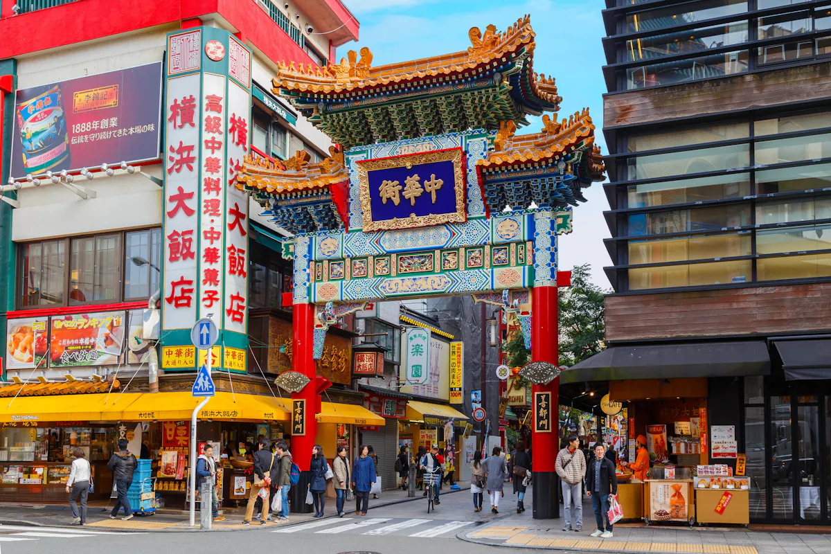 Yokohama's Chinatown Street scene in Yokohama Chinatown, featuring a colorful traditional Chinese gate with intricate designs. People walk and shop at various stores, including restaurants and souvenir shops. Bright signs with Chinese characters are prominently displayed on the buildings.