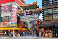 Street scene in Yokohama Chinatown, featuring a colorful traditional Chinese gate with intricate designs. People walk and shop at various stores, including restaurants and souvenir shops. Bright signs with Chinese characters are prominently displayed on the buildings.