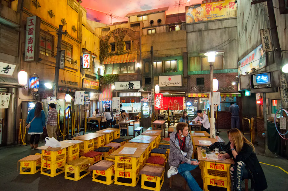 A vibrant indoor setting of a Japanese-themed street market. Stacks of yellow crates serve as makeshift tables and chairs. Various signs and lanterns hang from building facades. People are seated and chatting, giving the place a lively and bustling atmosphere.