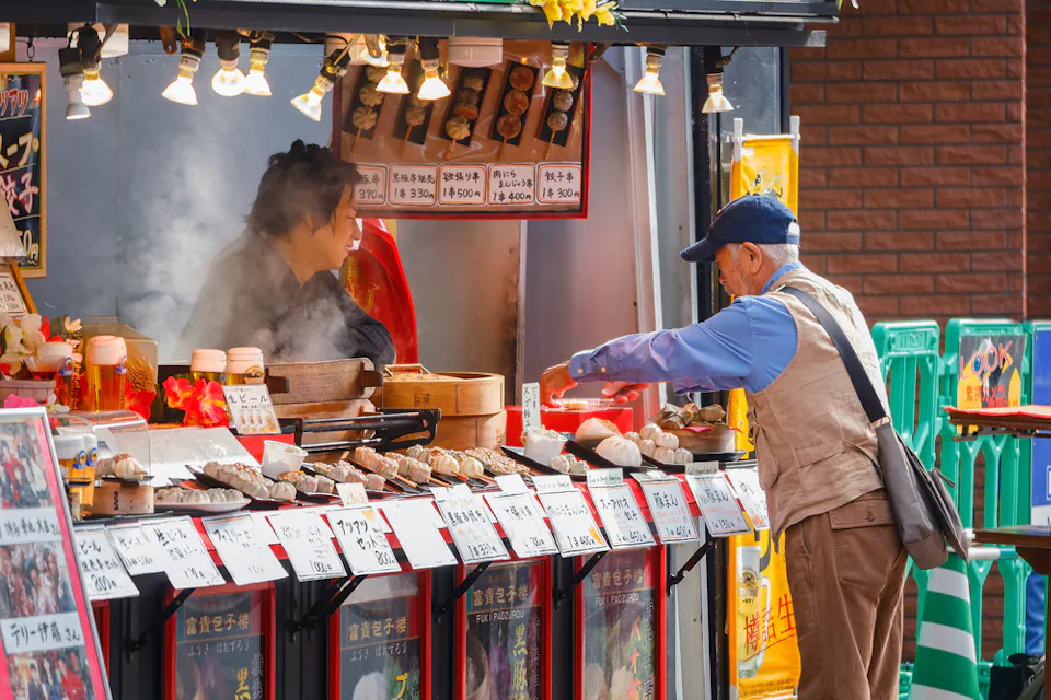 An elderly man wearing a blue cap and beige vest shops at a bustling food stall. The stall offers a variety of steamed and skewered foods, with steam rising from the dishes. A vendor behind the counter assists the man. Bright signs and menus are displayed on the stall.