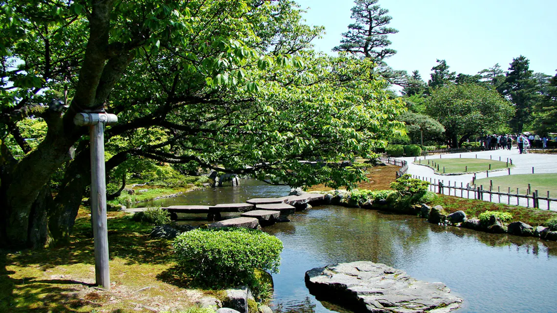 一个宁静的日本花园，特色是一个宁静的池塘，铺有石板路，郁郁葱葱的绿树，以及一棵枝繁叶茂的大树。背景中可以看到游客在探索花园的小径。