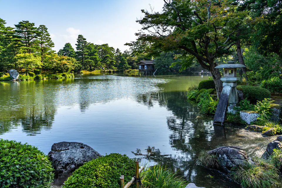 一个宁静的日本花园，中央有一个宁静的池塘，四周环绕着郁郁葱葱的绿树。一座石灯笼矗立在水边，一个小型传统木结构建筑在池塘对面可见。整个场景宁静，水面上倒映着树木的影子。