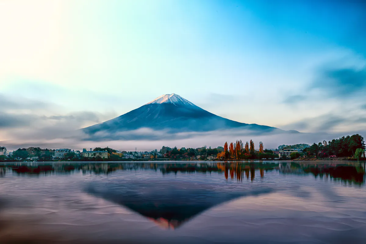 Mount Fuji A serene landscape features Mount Fuji at sunrise, its snow-capped peak silhouetted against a vibrant blue and orange sky. The mountain's reflection shimmers in calm lake waters, bordered by autumn foliage and misty, low-lying clouds.