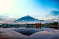 A serene landscape features Mount Fuji at sunrise, its snow-capped peak silhouetted against a vibrant blue and orange sky. The mountain's reflection shimmers in calm lake waters, bordered by autumn foliage and misty, low-lying clouds.