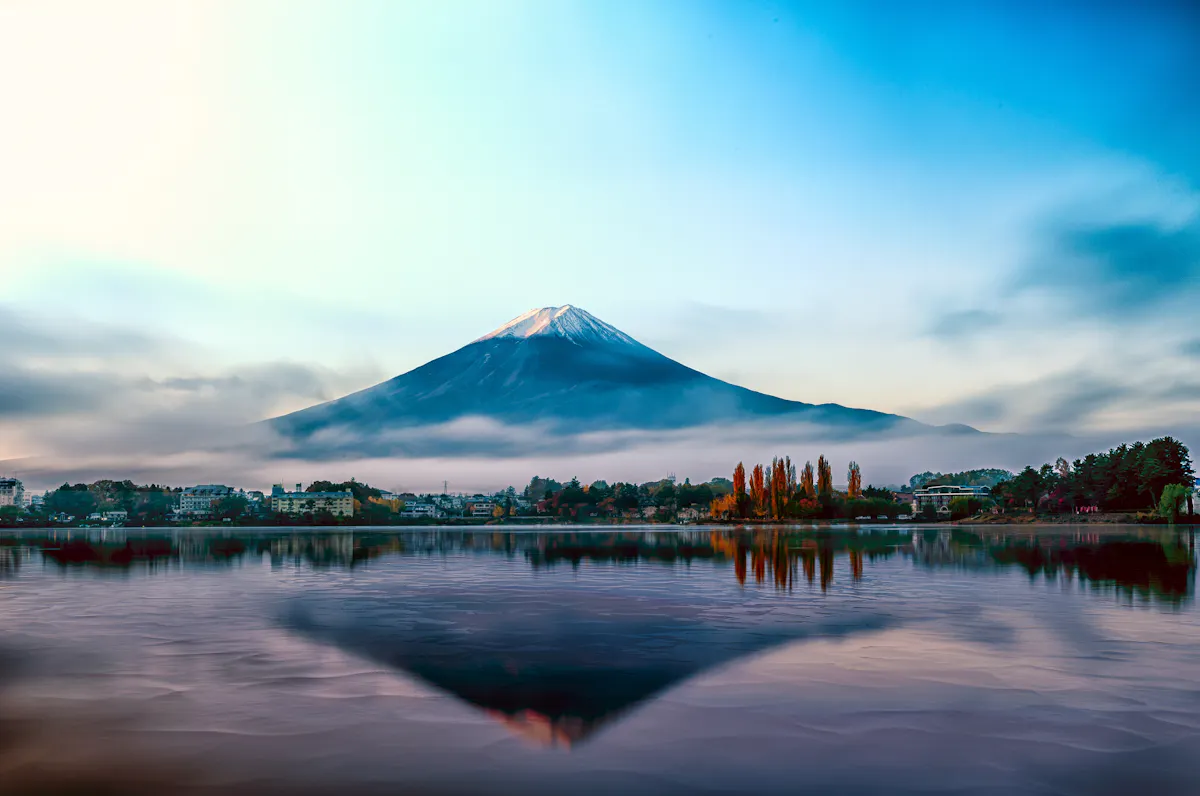 A serene landscape features Mount Fuji at sunrise, its snow-capped peak silhouetted against a vibrant blue and orange sky. The mountain's reflection shimmers in calm lake waters, bordered by autumn foliage and misty, low-lying clouds.