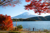 Mount Fuji with a snow-capped peak is seen in the background under a clear blue sky. In the foreground, vibrant red autumn leaves frame the scene. A serene lake rests between the mountain and the viewer, and a partially visible road borders the lake.