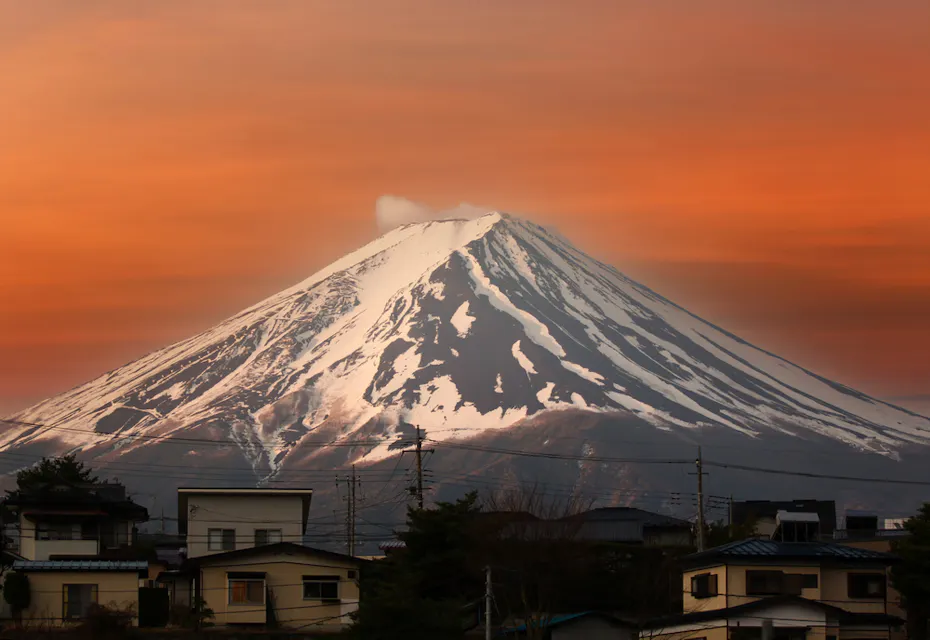 在日落时分，富士山的壮丽景色。白雪覆盖的山顶与生动的橙色天空形成了强烈对比。前景是一小片社区，分布着各种房屋和建筑。