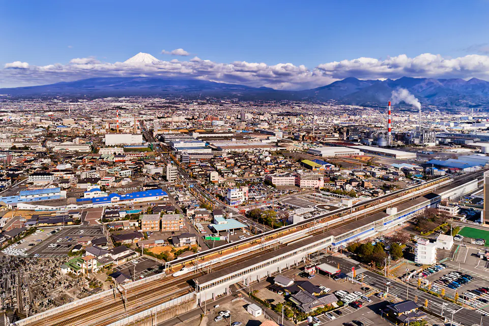 鸟瞰日本城市，建筑、道路和铁路交织。富士山在背景中部分被云层覆盖。城市景观包括住宅区、工业建筑和高架轨道上的高速列车。