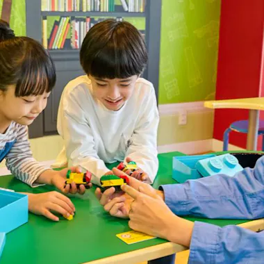 Tokyo Legoland Discovery Center Two children and an adult sit at a green table, smiling and playing with colorful LEGO bricks, with a bookshelf and bright green walls in the background.